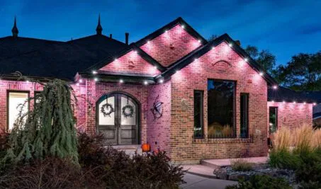A brick house at dusk with glowing pink and white uplighting along the roofline, arched double doors, two wreaths, tall grasses, shrubs, and a small pumpkin on the porch. The sky is deep blue.