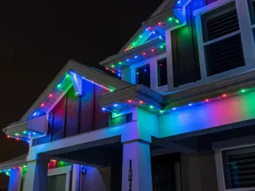 A house at night is decorated with colorful LED holiday lights along the roof and eaves, glowing in red, green, and blue. The house number 137 is visible on a white column.