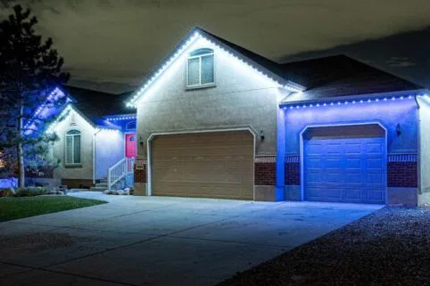 A suburban house at night with its garage doors closed, illuminated by rows of blue and white LED lights outlining the roof and eaves. The driveway is empty, and a tree is visible on the left side.