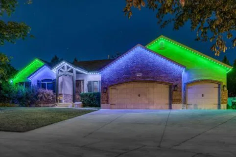 A brick house at night is decorated with bright green and white lights along the roofline. The garage doors are closed, and the lawn and driveway are visible in the foreground. Trees frame the scene.