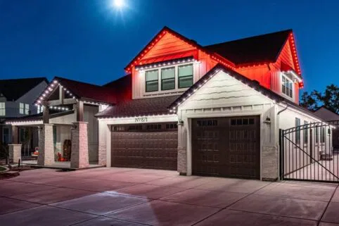 A modern house at night with red and white lights outlining the roof, three brown garage doors, a gated driveway, and a bright moon shining in the clear sky.
