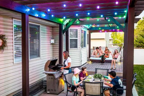 People enjoy a backyard gathering; one person grills food under colorful string lights, while others sit at a table eating, and a group relaxes on a raised deck nearby, with trees and a fence in the background.