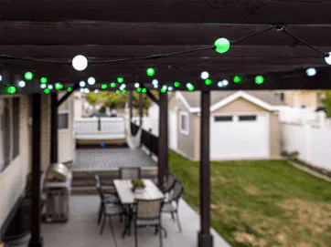 String lights with green and white bulbs hang from a wooden patio pergola above an outdoor dining table and chairs. In the background, there is a backyard with grass and a detached garage.