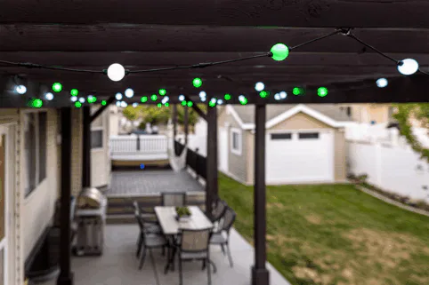 String lights with green and white bulbs hang from a wooden patio pergola above an outdoor dining table and chairs. In the background, there is a backyard with grass and a detached garage.