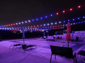 A swimming pool at night with red, white, and blue string lights hanging overhead. Lounge chairs and closed umbrellas surround the pool, and stone walls line the background. The area is empty and softly illuminated.