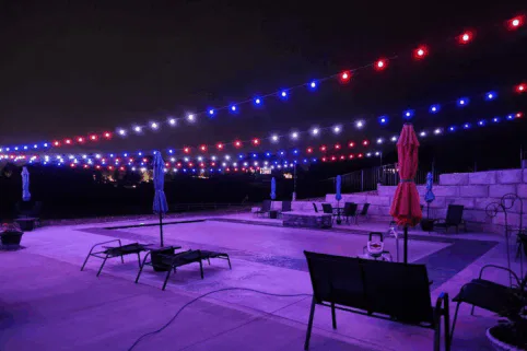 A swimming pool at night with red, white, and blue string lights hanging overhead. Lounge chairs and closed umbrellas surround the pool, and stone walls line the background. The area is empty and softly illuminated.