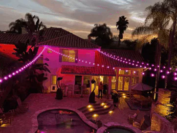 A backyard at dusk with a pool, patio furniture, and tropical plants. Purple string lights are draped across the area, illuminating the house and outdoor space with a warm, festive glow.