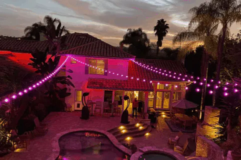 A backyard at dusk with a pool, patio furniture, and tropical plants. Purple string lights are draped across the area, illuminating the house and outdoor space with a warm, festive glow.
