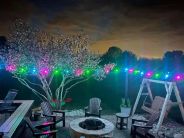 A backyard patio at night with a fire pit surrounded by chairs, a wooden swing, and a blooming tree illuminated by colorful string lights. The sky is dark with clouds and silhouetted trees in the background.