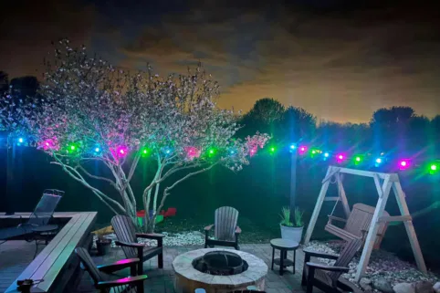 A backyard patio at night with a fire pit surrounded by chairs, a wooden swing, and a blooming tree illuminated by colorful string lights. The sky is dark with clouds and silhouetted trees in the background.