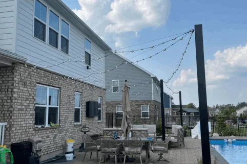 A backyard patio next to a brick house, featuring outdoor furniture, a table with an umbrella, string lights overhead, and a pool to the right. The scene is set in a suburban neighborhood under a partly cloudy sky.