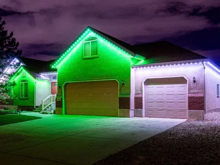 A suburban house at night with green and white LED lights outlining the roof, illuminating the driveway and garage, under a cloudy, purple sky.