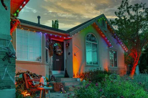 A house decorated for Halloween with colorful lights, fake spider webs, large spider props, and a skeleton seated on a chair by the front steps at dusk. Orange and green lights illuminate the scene.