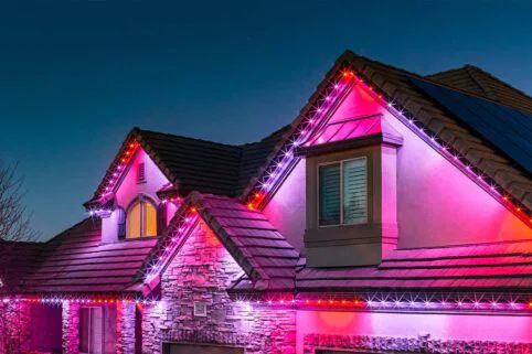 A house with stone and stucco exterior is decorated with red, purple, and white LED lights along the roofline, glowing brightly at dusk against a clear evening sky.