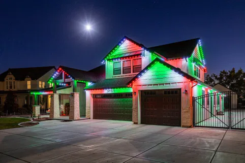 A two-story house at night is decorated with red, green, and white holiday lights along the roof. The sky is clear with a bright full moon shining above. The driveway and surrounding area are well-lit.