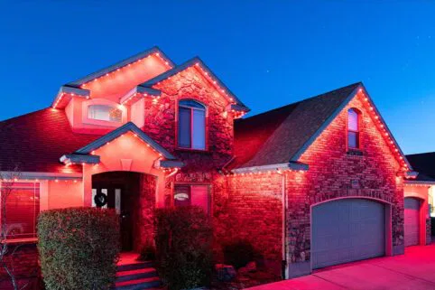 A two-story brick and stone house is decorated with red holiday lights outlining the roof and windows, glowing brightly at dusk under a clear blue sky.