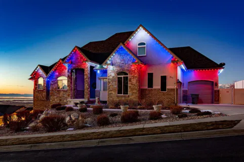 A house with stone and brick exterior is decorated with red, white, and blue lights along the roofline at dusk. The sky is clear, and landscaping with rocks and shrubs surrounds the home.