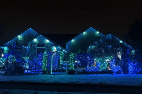 A house and yard decorated with blue and green holiday lights at night, with snow covering the ground and bushes, creating a festive winter scene.