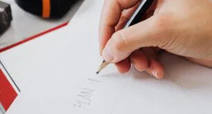 Attached image file A person sketches on white paper with a pencil at a desk, near a red notebook, a black tape measure, and a laptop.