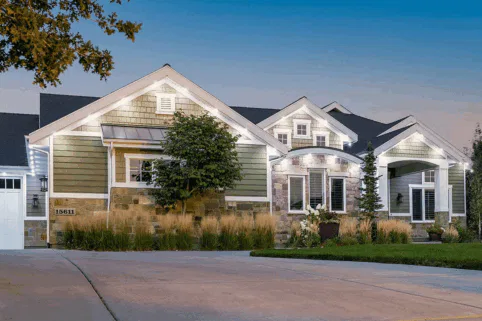 A well-lit suburban house with green and stone exterior, multiple gables, a manicured lawn, and a driveway at dusk. Shrubs and small trees decorate the front yard.