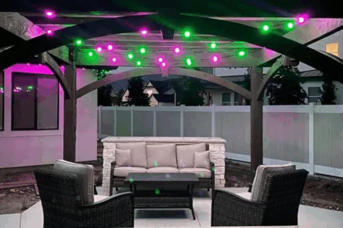 A backyard patio seating area with a couch, two chairs, and a table under a pergola. Purple and green string lights are hung across the pergola beams, illuminating the outdoor space.