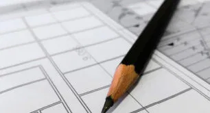Attached image file A sharpened black pencil rests on top of architectural blueprints, which display technical drawings and plans with various lines, shapes, and measurements.