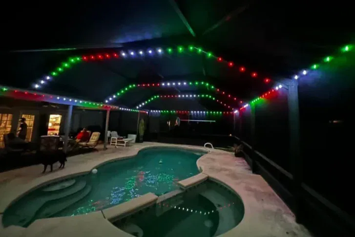 A backyard pool at night is lit by red, green, and white string lights hanging from a covered patio roof. The pool area has lounge chairs, and a few people and a dog are visible near the illuminated house.
