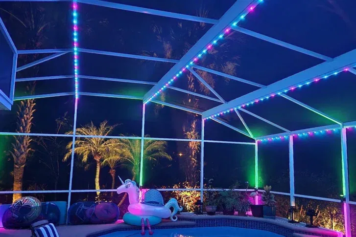 A screened patio at night is lit with colorful LED lights along the frame. There are palm trees outside, inflatable pool floats including a unicorn, and various poolside items around a small pool.