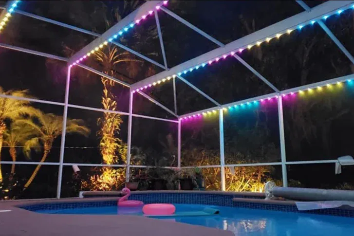 A swimming pool at night with colorful string lights along the screened enclosure, palm trees outside, and pool floats including a pink flamingo and a round float in the water.