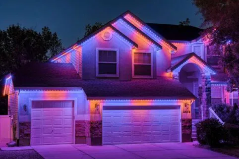 A two-story house at dusk is decorated with vibrant purple, pink, and blue LED lights outlining its roof, garage, and entryway, creating a colorful and festive appearance against the night sky.