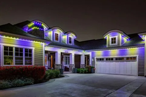 A modern house at night with purple, blue, and white LED lights outlining the roof. The driveway and front yard are visible, and the garage door is closed.