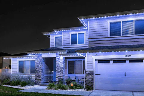 A modern two-story house at night is decorated with white string lights along the roofline, creating a festive, welcoming appearance. The house has stone pillars, large windows, and a well-kept front yard.