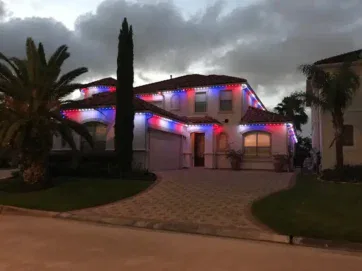 A two-story house is decorated with red, white, and blue lights along the roofline. Palm trees and shrubs are in the front yard, and the sky is cloudy at dusk.