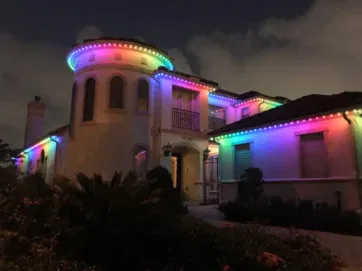 A two-story house is decorated with multi-colored string lights along the roofline, glowing in shades of red, green, blue, and purple at night. Cloudy sky and plants are visible in the foreground.
