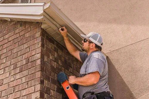A man wearing a gray shirt, cap, and sunglasses stands on an orange ladder, inspecting or repairing the gutter of a brick house under a partly cloudy sky.