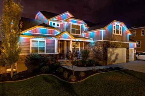 Two-story house at night with stone and wood siding, illuminated by colorful, multicolored LED lights outlining the roof and windows. The yard is landscaped with trees and shrubs.