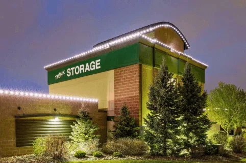 A self-storage facility building with a green sign reading “THINK STORAGE,” surrounded by pine trees and shrubs. The building is decorated with white string lights along the roofline, and the sky is softly lit at dusk.