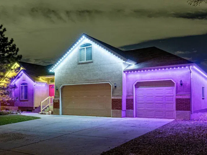 A house at night with purple and white LED strip lights outlining the roof, garage doors, and entrance; the driveway and yard are visible in the foreground.